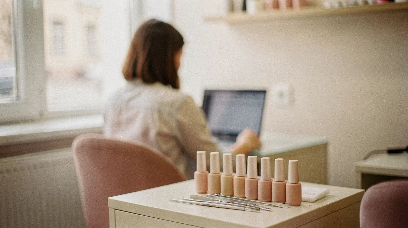 Kobieta pracuje przy laptopie, na pierwszym planie rząd lakierów i narzędzia do manicure