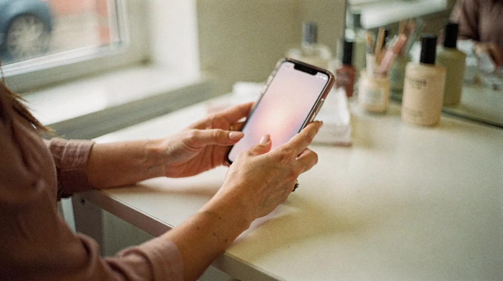 Dłonie trzymają smartfon z rozmytym ekranem przy toaletce z kosmetykami w salonie